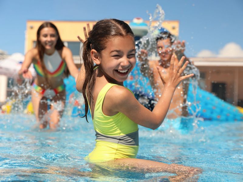 Smiling girl in a yellow swimsuit splashing water in a pool with two kids playing in the background.