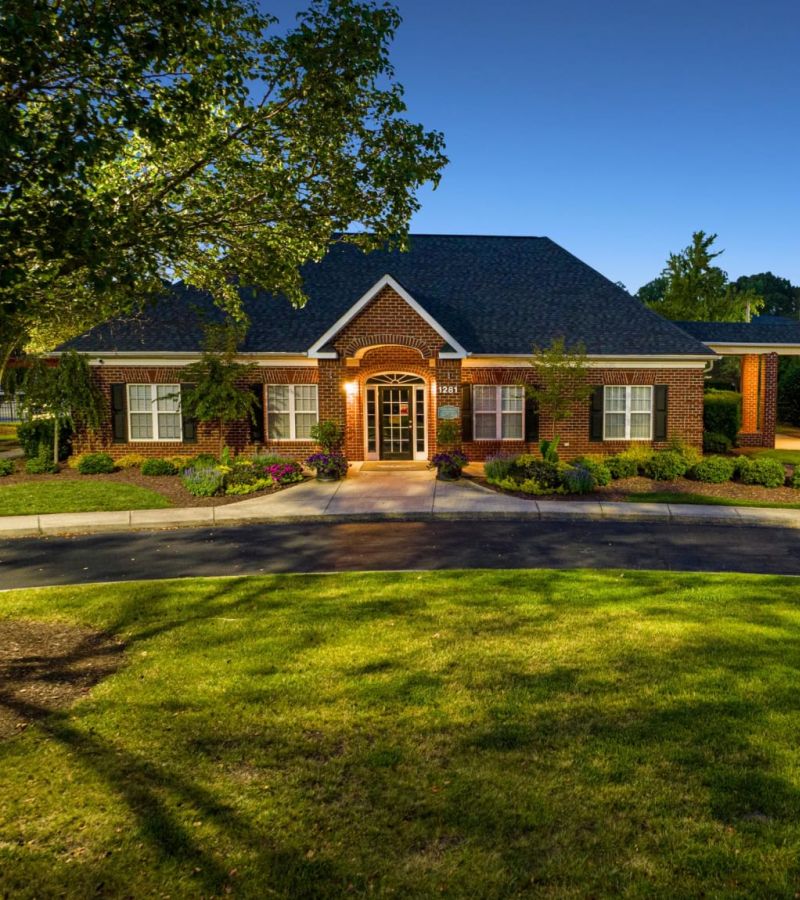 Brick house with lit entrance at dusk, surrounded by trees, bushes, and a well-manicured lawn.