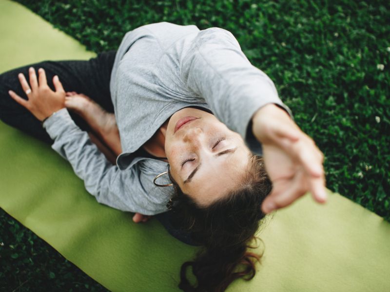 Woman stretching on a yoga mat outdoors, eyes closed, reaching one arm forward over her head.