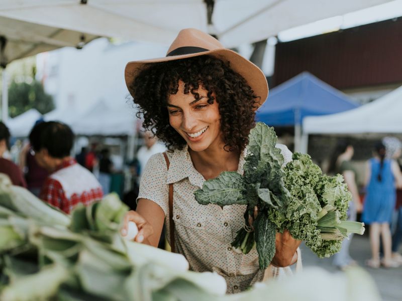Woman smiling while shopping for leafy greens at an outdoor market, holding fresh kale in her hand.