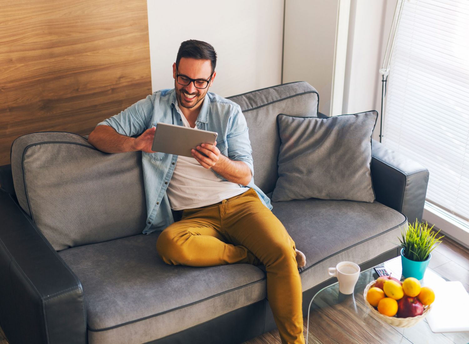 Smiling man sitting on a gray couch using a tablet, with coffee and fruit on the table nearby.