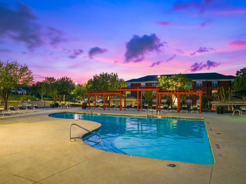 Outdoor pool with lounge chairs and pergolas at sunset, with trees and a building in the background.