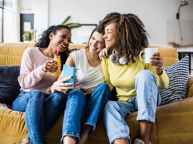 Three women sitting on a couch, smiling and taking a selfie together, each holding a drink.