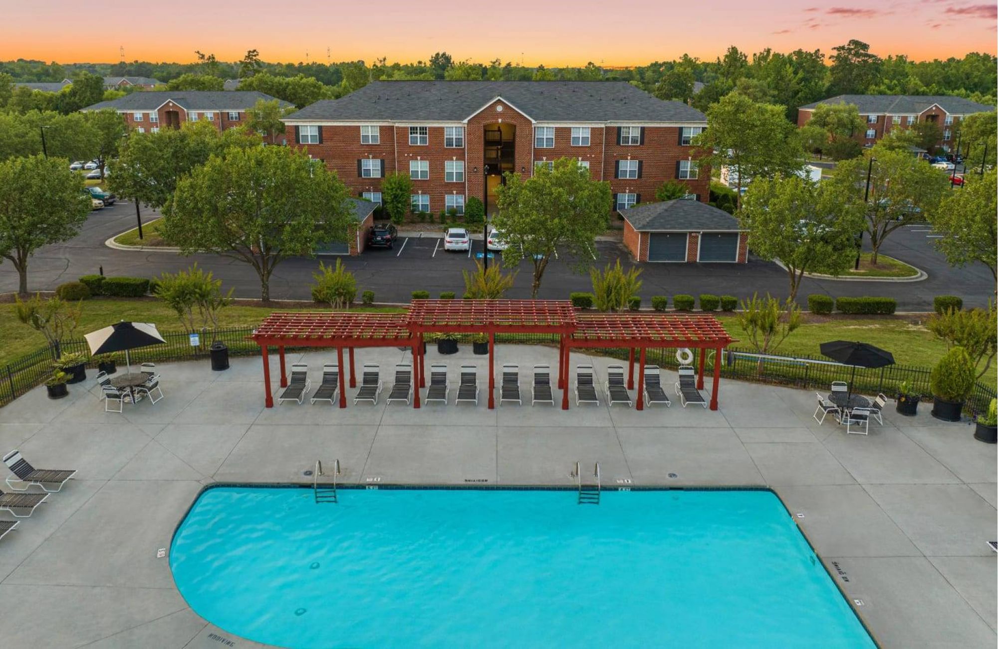 Outdoor pool area with lounge chairs, pergola, and brick apartment buildings at sunset.