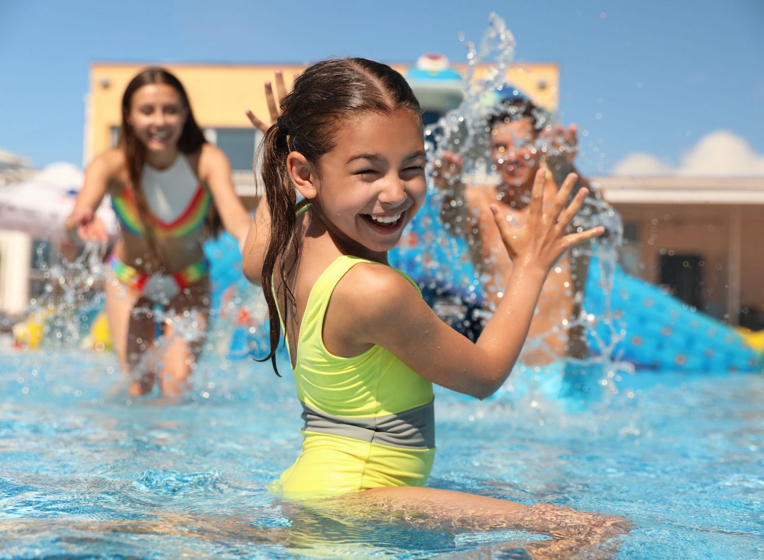 Smiling girl in a yellow swimsuit splashing water in a pool with two kids playing in the background.