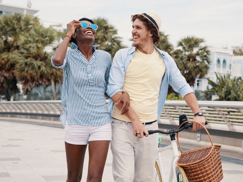 A smiling couple walks outdoors with a bicycle, enjoying a sunny day near palm trees and buildings.