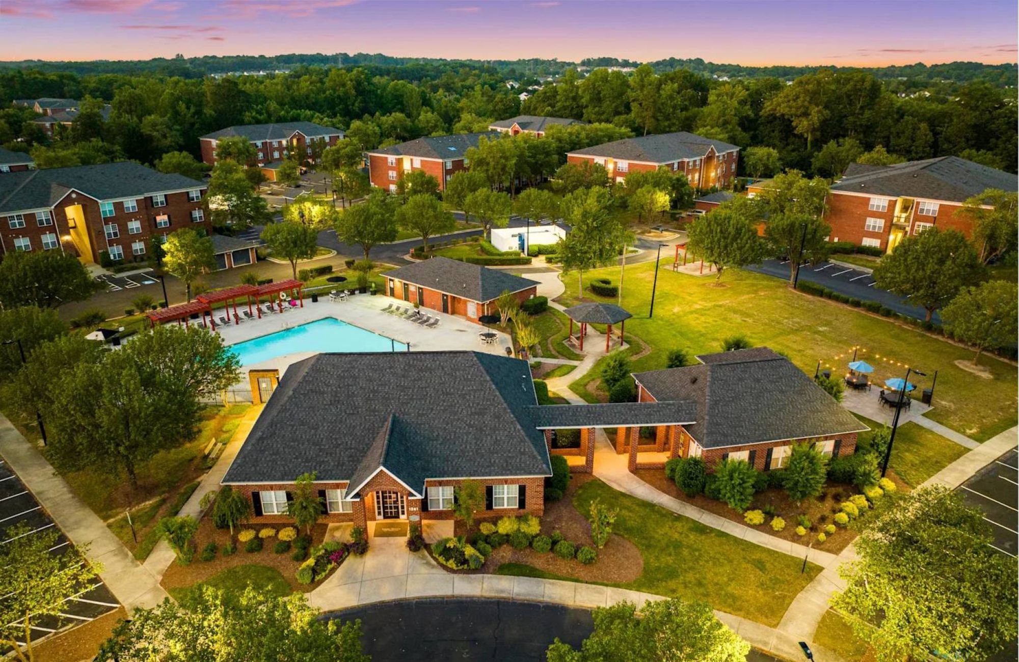 Aerial view of a residential complex with a pool, clubhouse, trees, and several apartment buildings at sunset.