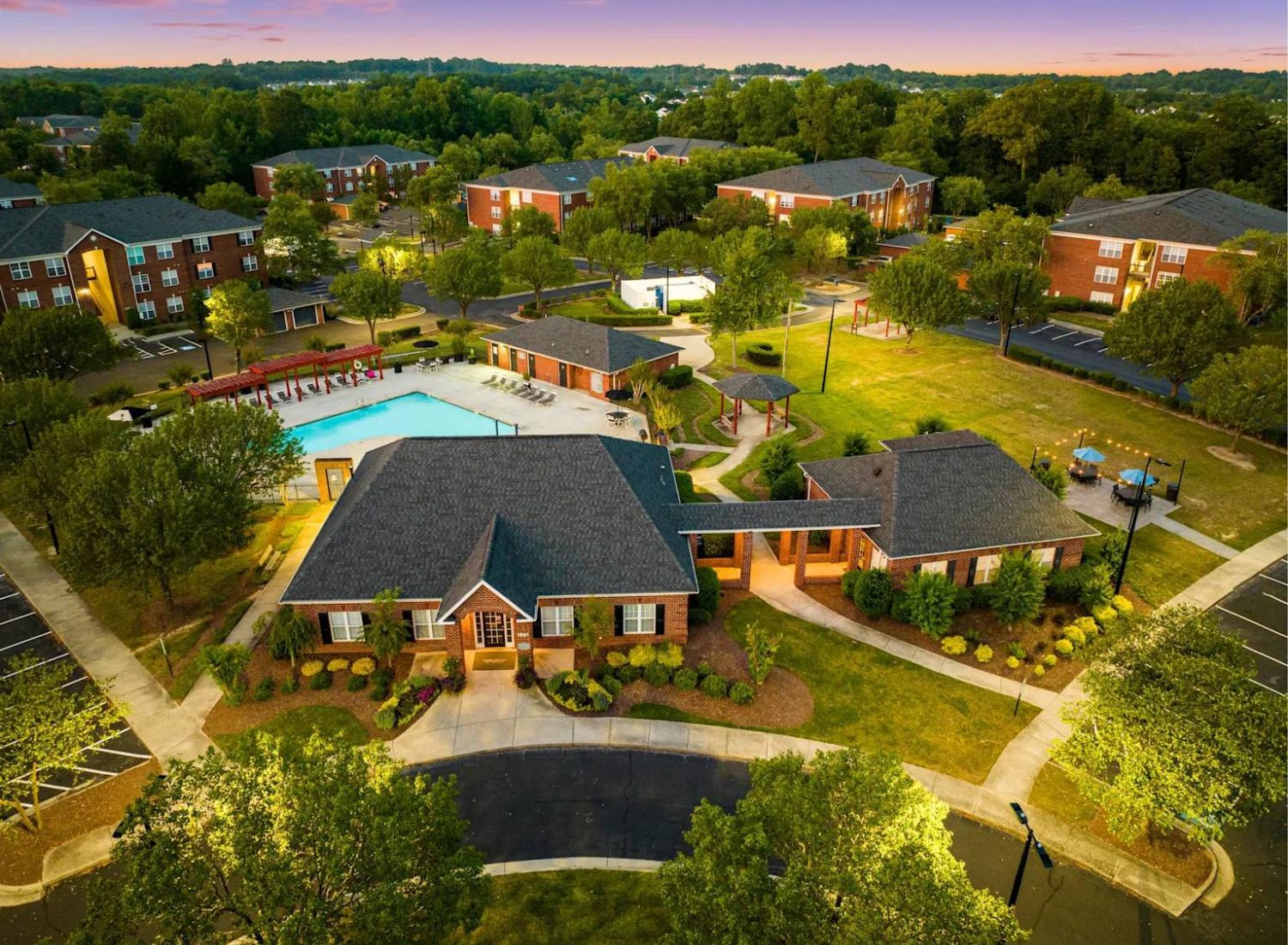 Aerial view of a residential complex with a pool, clubhouse, trees, and several apartment buildings at sunset.